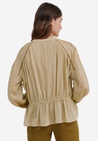 Blusa beige chiaro con motivo testurizzato, vita arricciata e maniche a palloncino. Presenta una delicata cucitura lungo le spalle.