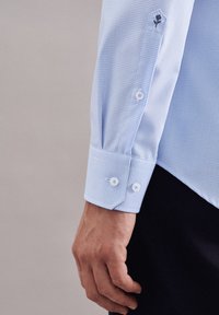 Lyseblå ternet skjortemanchet med hvide knapper på en persons håndled og hånd mod en neutral baggrund.