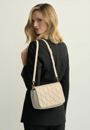 Blondynka w czarnym blezerze, niosąca kremową pikowaną torebkę na ramię z regulowanym paskiem, spoglądająca przez ramię na jednolitym tle.