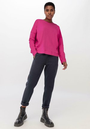 Magenta lange mouwen top en donkere broek. De broek heeft omgezoomde broekspijpen. Zwarte enkellaarsjes met gestructureerde zolen. Zachte stofstructuren zichtbaar.
