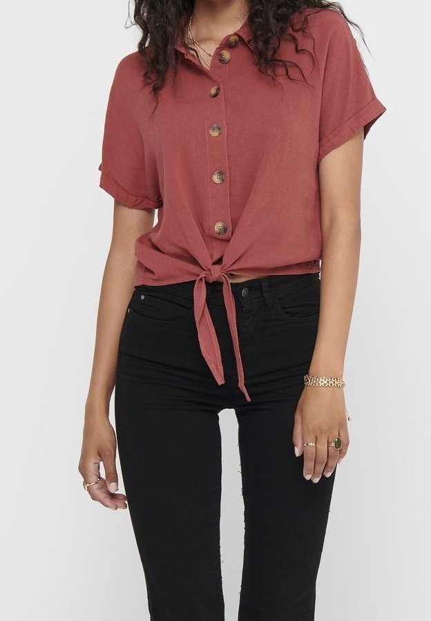 Femme portant une chemise à manches courtes couleur rouille nouée à la taille et un pantalon noir taille haute, avec des bijoux aux mains.