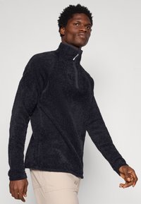 Svart fleece-pullov med hög krage och delvis dragkedja, med en mjuk, texturerad yta och långa ärmar. Bärs över ljusa byxor.