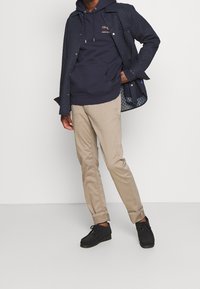 Chaqueta de napa azul marino sobre una sudadera con capucha azul marino con un logo dorado, combinada con pantalones beiges y zapatos negros. El atuendo presenta elementos de diseño simples.