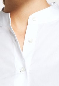 Chemise blanche à col avec patte de boutonnage et deux boutons visibles, en tissu lisse. Présente un col mao et un design épuré et minimaliste.