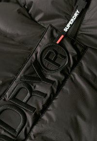 Svart puffjacka med texturerat tyg, featuring stor broderad "DRY"-logotyp, kontrasterande etikett märkt "SUPERDRY", och polerad finish.