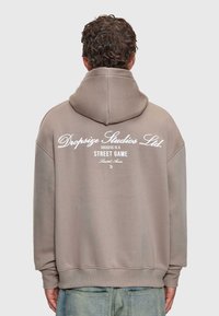 Mann mit lockigem Haar, der einen taupefarbenen Hoodie mit weißem Text "Dropsize Studios Ltd. Erfolg ist ein Straßen-Spiel mit begrenztem Zugang" auf dem Rücken trägt.