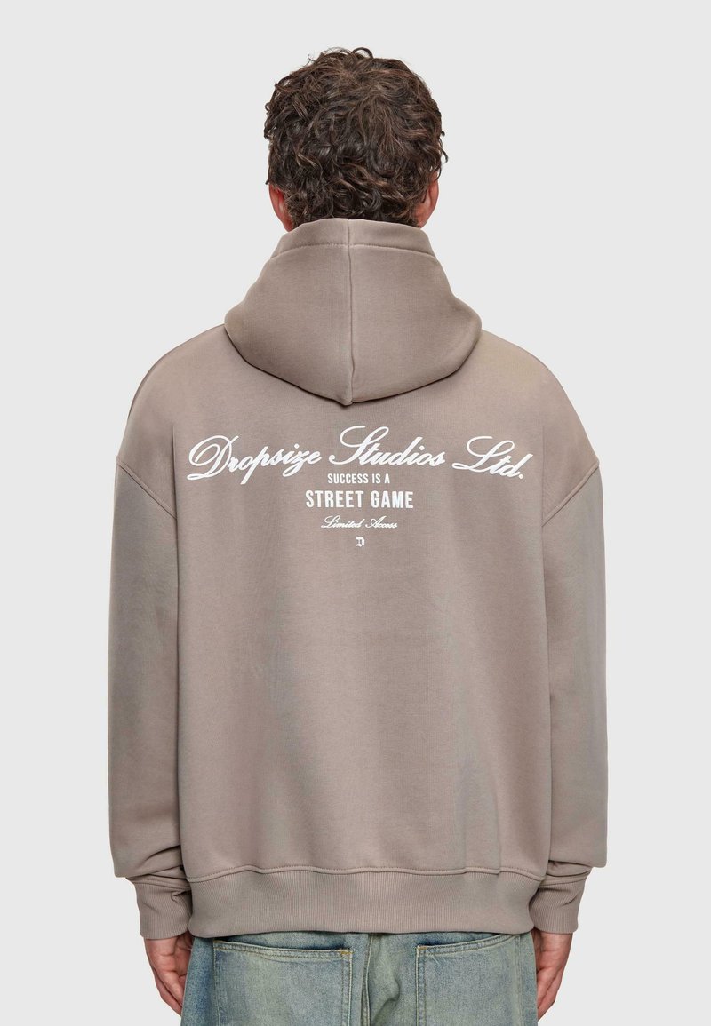 Mann mit lockigem Haar, der einen taupefarbenen Hoodie mit weißem Text "Dropsize Studios Ltd. Erfolg ist ein Straßen-Spiel mit begrenztem Zugang" auf dem Rücken trägt.
