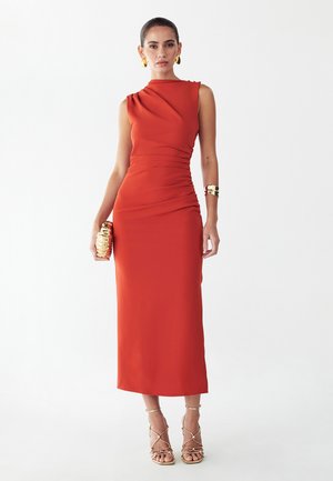 Vrouw draagt een mouwloze, strak zittende midi-jurk in verbrand oranje met geplooide details, gouden oorbellen, armband, clutch en sandalen met hoge hakken en bandjes.
