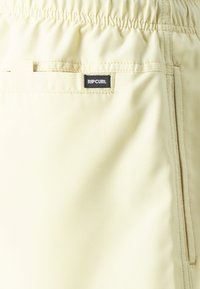 Shorts de bain jaune clair avec une taille élastique et une étiquette "RIP CURL" noire sur la poche arrière. Texture lisse, design minimal.