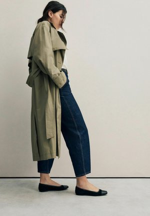 Khaki trenchcoat, witte shirt, donkerblauwe jeans en zwarte ballerina's. De jas heeft een ceintuur in de taille en een dubbele knoopsluiting.