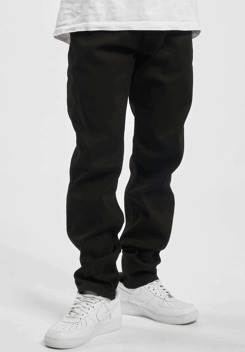 Person med svarte bukser og hvite sneakers står på en hvit bakgrunn, beskåret over livet.