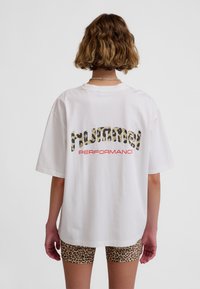 Hvid oversized t-shirt med leopardprinttekst på bagsiden, hvor "PERFORMANCE" står med rødt, kombineret med leopardprintshorts.