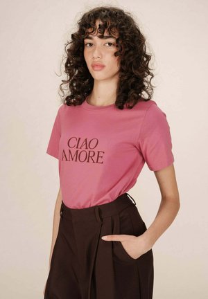 Femme aux cheveux bouclés et foncés portant une chemise rose avec le texte "CIAO AMORE" et un pantalon noir taille haute, avec une main dans la poche.