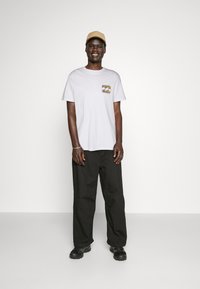 Bílá trička s barevným designem kapsy, spárovaná s černými volnými kalhotami a béžovou čepicí. Černé boty dokončují celý outfit.