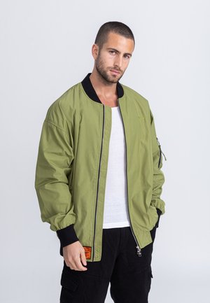 Oliwkowa kurtka bombowca z czarnym kołnierzem, zamek błyskawiczny i boczne kieszenie. Posiada elastyczne mankiety oraz pomarańczową metkę na dolnym brzegu.