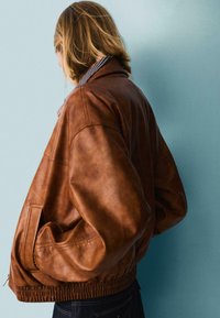 Giacca bomber in pelle marrone con orlo a coste, vestibilità oversize, tasche laterali e colletto rigato, su uno sfondo azzurro chiaro.