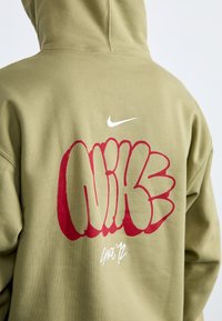 Olivinės spalvos džemperis su dideliu raudonu graffiti stiliaus "NIKE" logotipu ir baltu „swoosh“ simboliu. Po tekstu rašoma "Nuo '12."