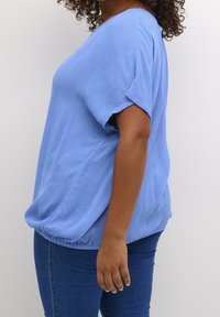 Top azul claro de tela texturizada y corte holgado. Tiene mangas cortas y un dobladillo fruncido. Combinado con jeans ajustados azules.