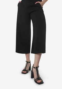 Zwarte wijdvallende culotte-broeken met zijzakken. Gepaard met zwarte sandalen met hielbandjes, voorzien van een transparante accent en een chunky hak.