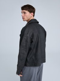 Giacca nera in ecopelle con colletto classico, polsini con cerniera e dettagli di cuciture visibili, indossata da una persona di profilo.