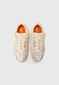 Nike-sko i beige og grå mesh, med kremfargede snørebånd, oransje detaljer, og en fremtredende logo på tungen og siden.
