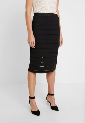 Kvinde iført en sort knælange nederdel med horisontale udskårne striber og sorte højhælede sko med ankelrem, kombineret med en hvid blondebluse.