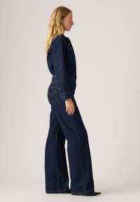 Macacão de denim azul escuro, com um topo ajustado de mangas longas e calças de perna larga. Detalhe de costura notável e bolsos tradicionais.