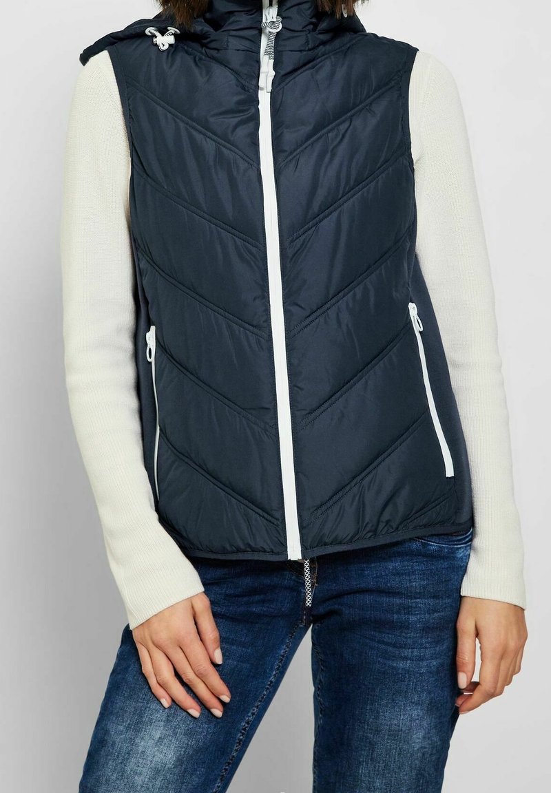 Námořnický modrý prošívaný vest, s chevronovým prošíváním, bílým zipem a nastavitelnou kapucí, nošený přes krémový dlouhý rukáv, spárovaný s džíny.