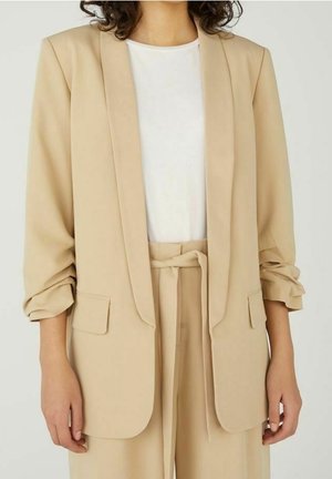 Personne portant un blazer beige cintré avec les manches retroussées sur une chemise blanche et un pantalon taille haute assorti avec ceinture nouée, debout devant un fond uni.
