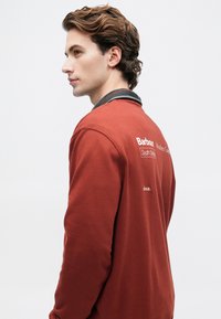 Νεαρός άνδρας με καστανά μαλλιά, φορώντας μια κολοκυτόχρωμη μπλούζα με λευκό κείμενο στην πλάτη, μπροστά από ένα απλοϊκό ανοιχτό φόντο.