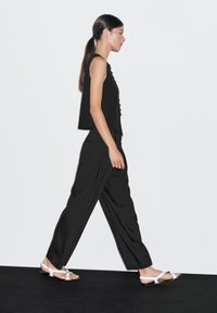 Schwarzes ärmelloses Oberteil mit Knöpfen vorne, weite schwarze Hose und weiße Sandalen. Model geht vor einem schlichten weißen Hintergrund.