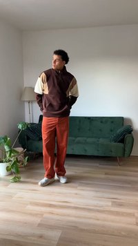 Καφέ και μπεζ fleece μπουφάν με χρώματα μπλοκ, πορτοκαλί παντελόνι από κοτλέ, άσπρα sneakers; στέκεται σε δωμάτιο με πράσινο καναπέ και φυτό.