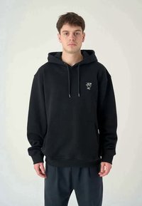 Young man wearing black hoodie with small white bird logo on chest, standing against plain light background.
