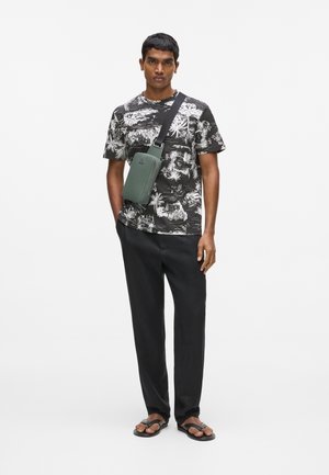 Mand iført sort og hvid t-shirt med tropisk print, sorte bukser, sorte sandaler og grøn crossbody-taske mod hvid baggrund.