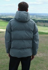 Jachetă puffer într-o nuanță verde mat, cu guler înalt și glugă, având un design matlasat și o textură netedă, mat.