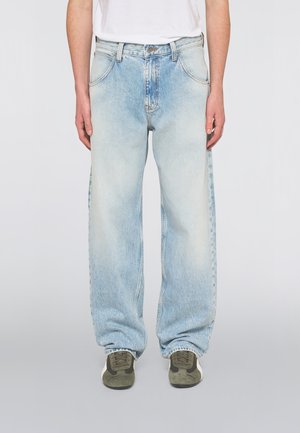 Person trägt hellblaue, ausgewaschene, weite Jeans, weißes Hemd und grüne Turnschuhe und steht vor einem einfarbigen Hintergrund.