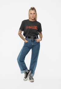 Dark gray t-shirt with red "Gorillaz" logo; high-waisted blue denim jeans; black and white sneakers; casual fit; tattoos visible on arms.