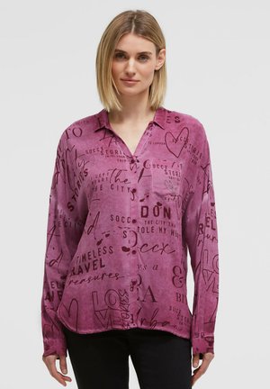 Frau trägt ein langärmeliges lila Hemd mit schwarzen abstrakten Text- und Herzdesigns, kombiniert mit schwarzen Hosen.