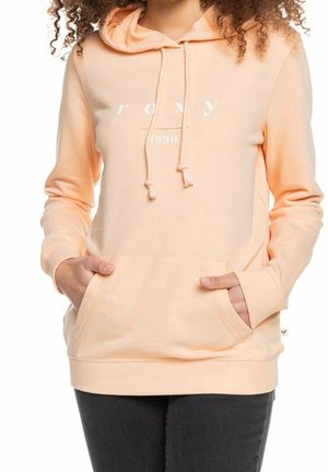 Kvinde iført ferskenfarvet Roxy hoodie med frontlomme, snore og teksten "roxy 1990", kombineret med mørke bukser.