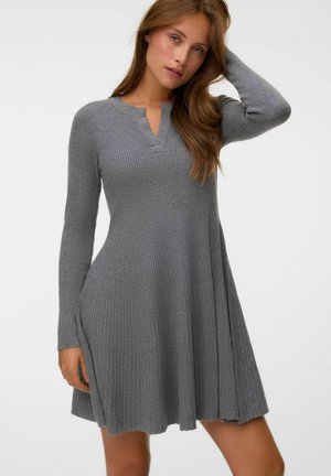 Femme portant une robe en maille grise côtelée à manches longues, avec un petit col en V, posant avec une main touchant ses cheveux.