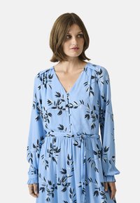 Blue long-sleeve dress with a V-neck, featuring a floral pattern in black and white, and a cinched waist with a tie. Buttons visible.