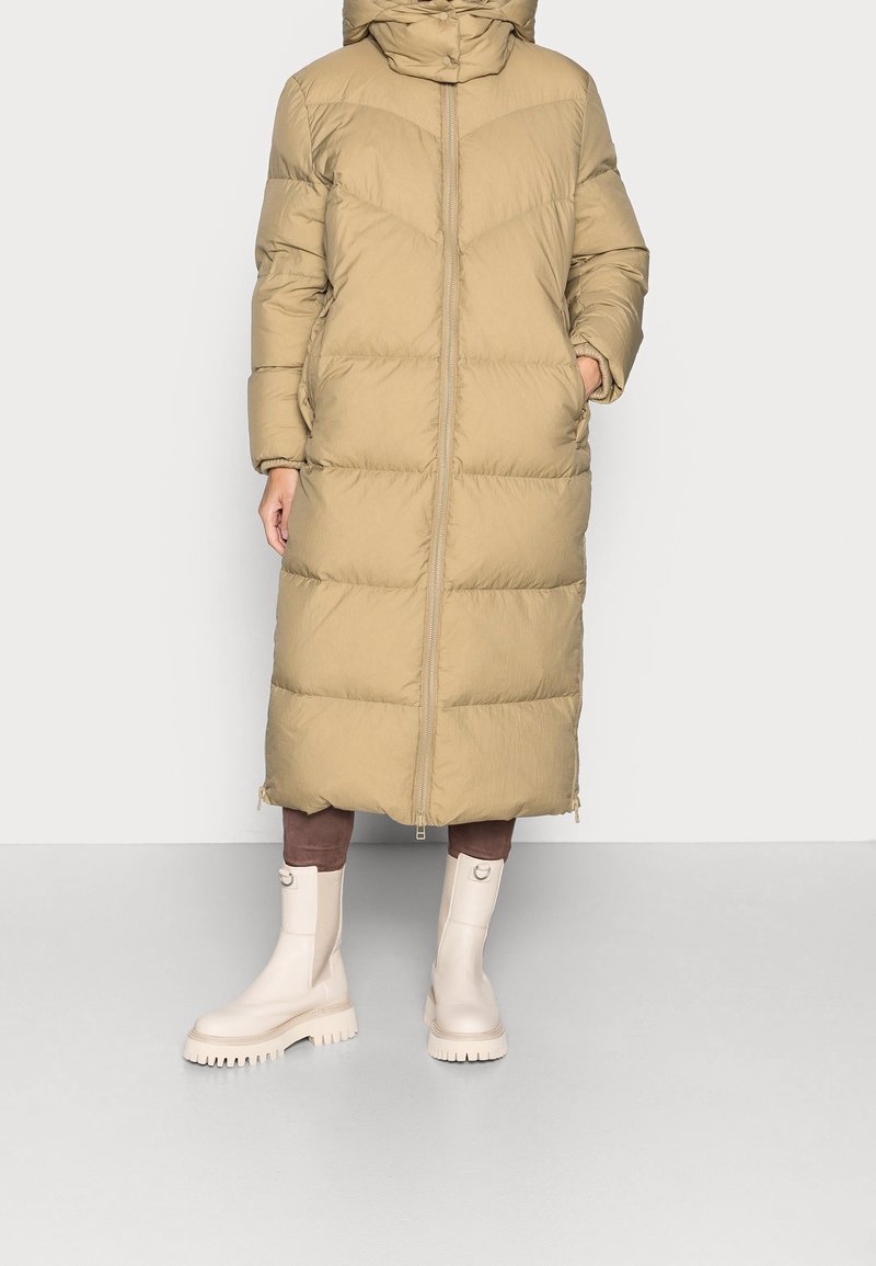 Abrigo acolchado beige texturizado con cremallera completa, capucha y largo hasta media pierna. Acompañado por botines color crema con suela gruesa.