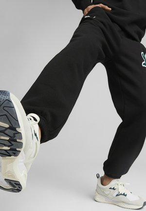Svart joggebukse med ribbestrikkede mansjetter, med et brodert grønt logo og sidelommer. Båret med hvite sko og grå yttersåle.