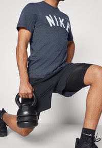 Tummansininen Nike t-paita, jossa on suuri valkoinen logo, yhdistettynä mustiin urheiluhousuihin ja mustiin lenkkareihin, pitäen mustaa kahvakuulaa.