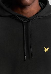 Felpa nera con cappuccio realizzata in tessuto liscio, dotata di tasca frontale e cordini. Un piccolo logo giallo a forma di aquila è posizionato sul petto.