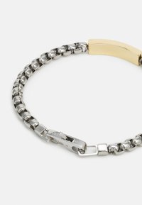 Pulseira de corrente em tom prateado com elos de metal polido, um detalhe dourado e um fecho com gravação da marca.
