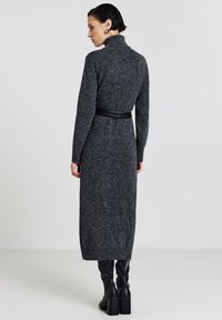 Robe en tricot grise longue avec un col montant et une taille ceinturée. Le mannequin porte des bottes à talons noires montantes jusqu'aux genoux. Texture lisse et coupe slim.