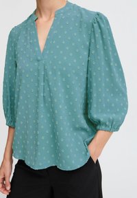 Blusa teal con scollatura a V, maniche a palloncino elasticizzate e un tessuto leggero e tessurizzato con piccoli motivi sparsi. Orlo ondulato.