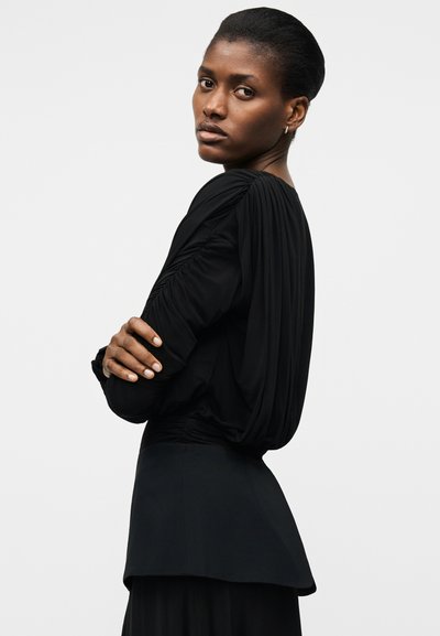 Kobieta o krótkich włosach, ubrana w czarną, marszczoną bluzkę z długim rękawem i czarną spódnicę, spoglądająca przez ramię z założonymi skrzyżowanymi ramionami.