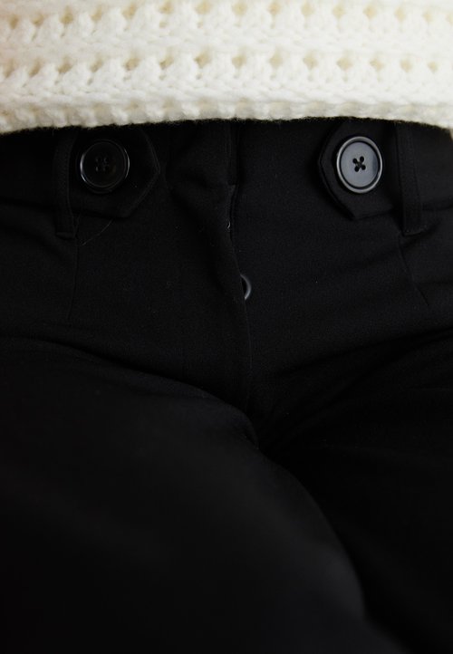Schwarze Hosen mit glatter Textur und zwei schwarzen Knöpfen an der Taille, teilweise von einem cremefarbenen Strickoberteil bedeckt.